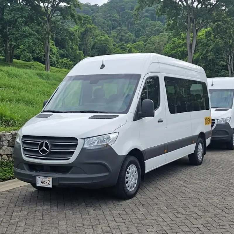 Line of modern white Mercedes-Benz Sprinter executive passenger vans parked on a paved road surrounded by lush tropical green hills in Costa Rica.